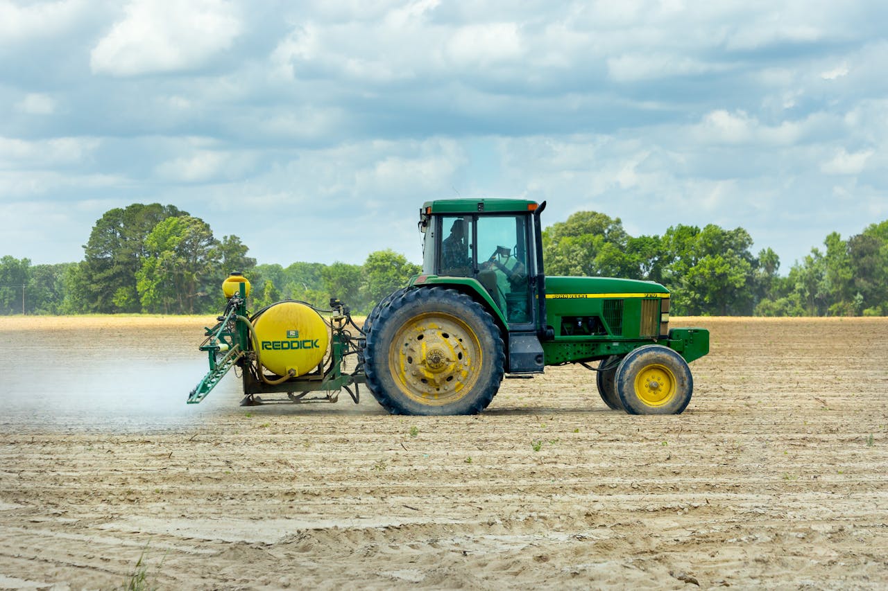about-us Green tractor with sprayer working in an open farm field under a cloudy sky.